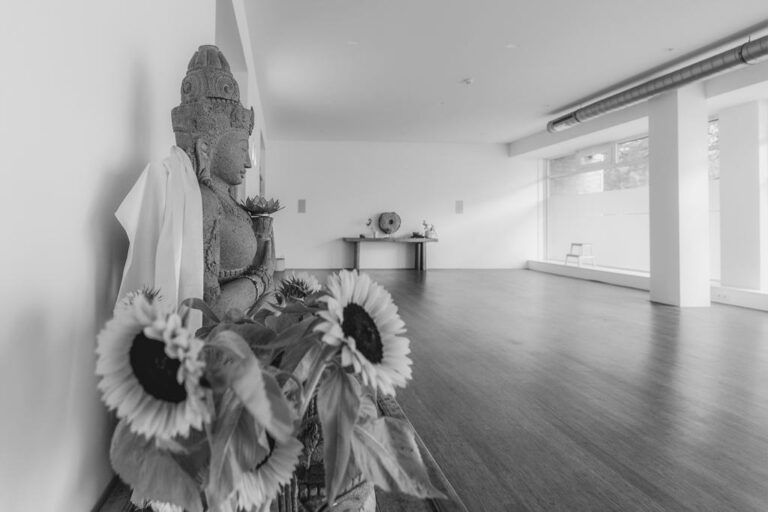 The Inner Embassy yoga studio: a calm, light-filled room with a wooden floor, a stone deity statue draped in cloth, and sunflowers by the window
