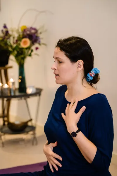 Irene Lähde in profile during a breathing practice, one hand resting on her chest, in a calm candlelit studio with fresh flowers